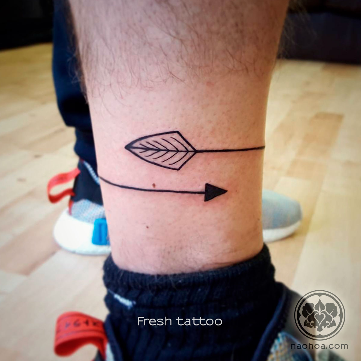 A tattoo of a bold, minimalist arrow that wraps around the client's leg. Designed and tattooed by Naomi Hoang at NAOHOA Luxury Bespoke Tattoos, Cardiff (Wales, UK).
