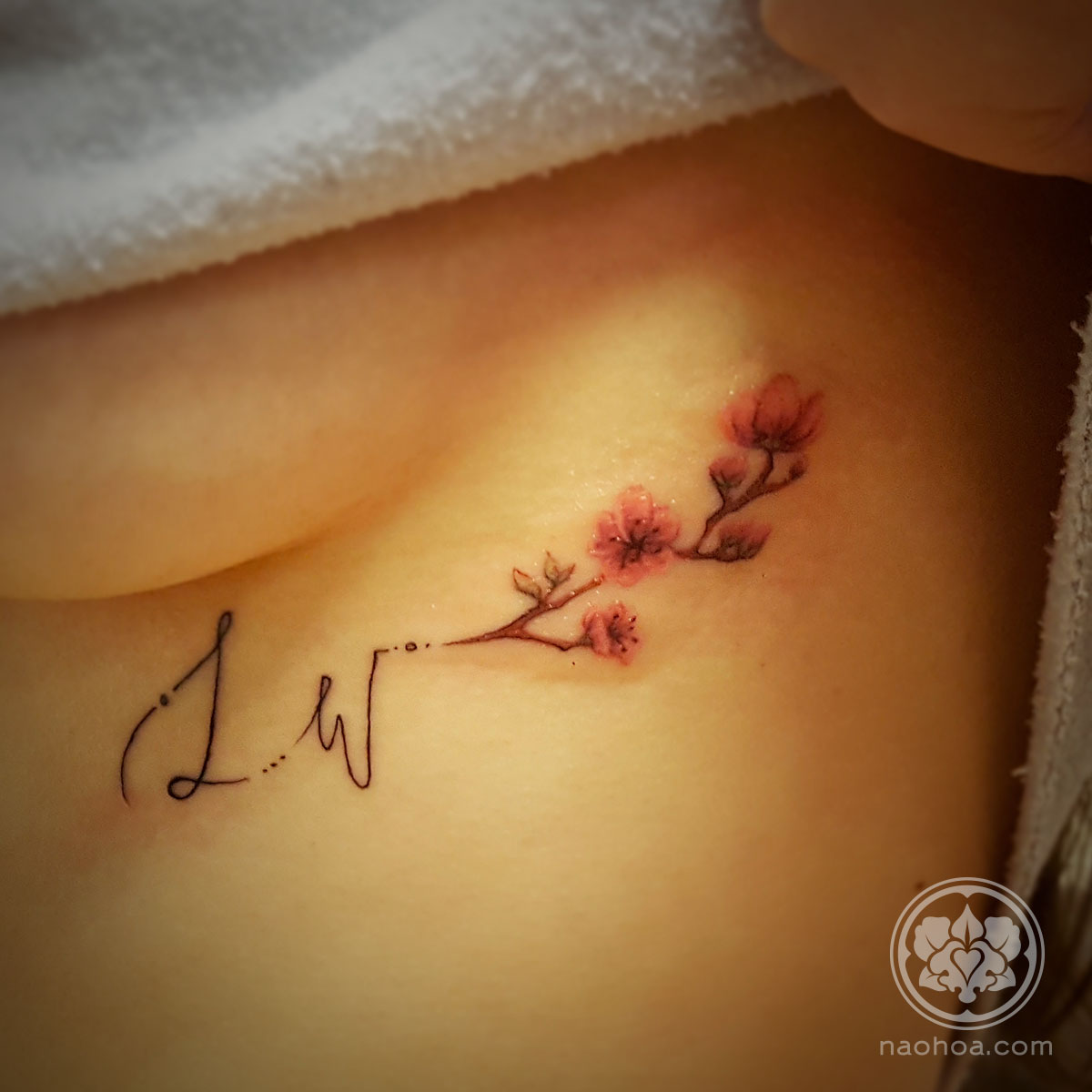 A delicate tattoo of the initials 'LW' with a branch of cherry blossoms, placed just under the woman's breast. Designed and tattooed by Naomi Hoang at NAOHOA Luxury Bespoke Tattoos, Cardiff (Wales, UK).