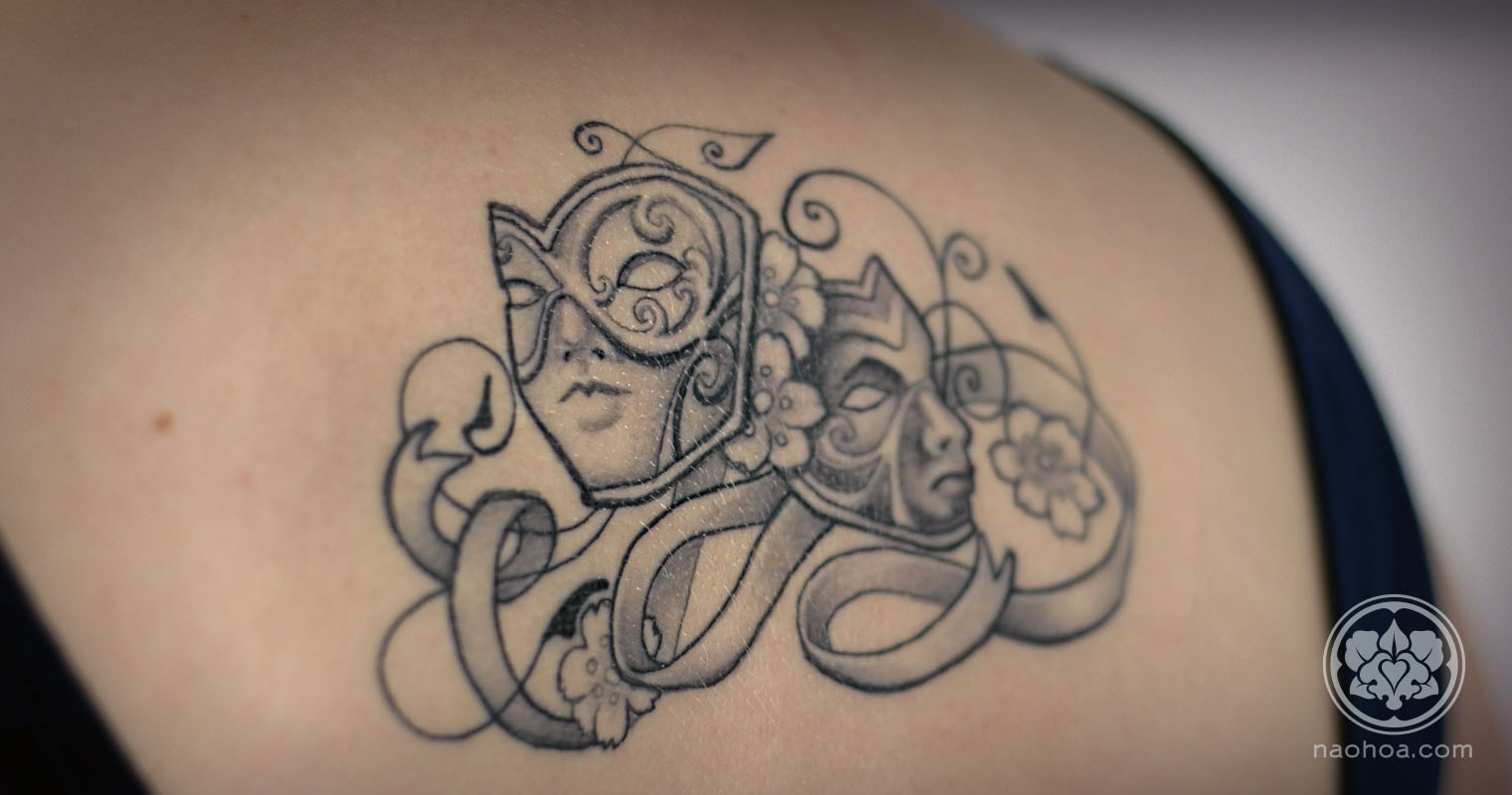 Black and white tattoo of two theatre masks, surrounded by flowers and swirling ribbons.